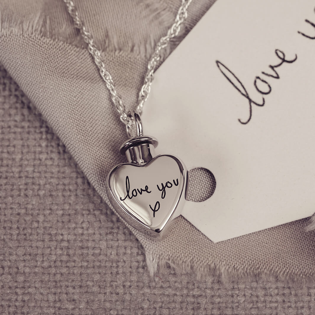 A silver, heart shaped self-fill urn necklace, on a silver chain, with a handwritten message of 'love you x' engraved into the front. Displayed on a neutral background