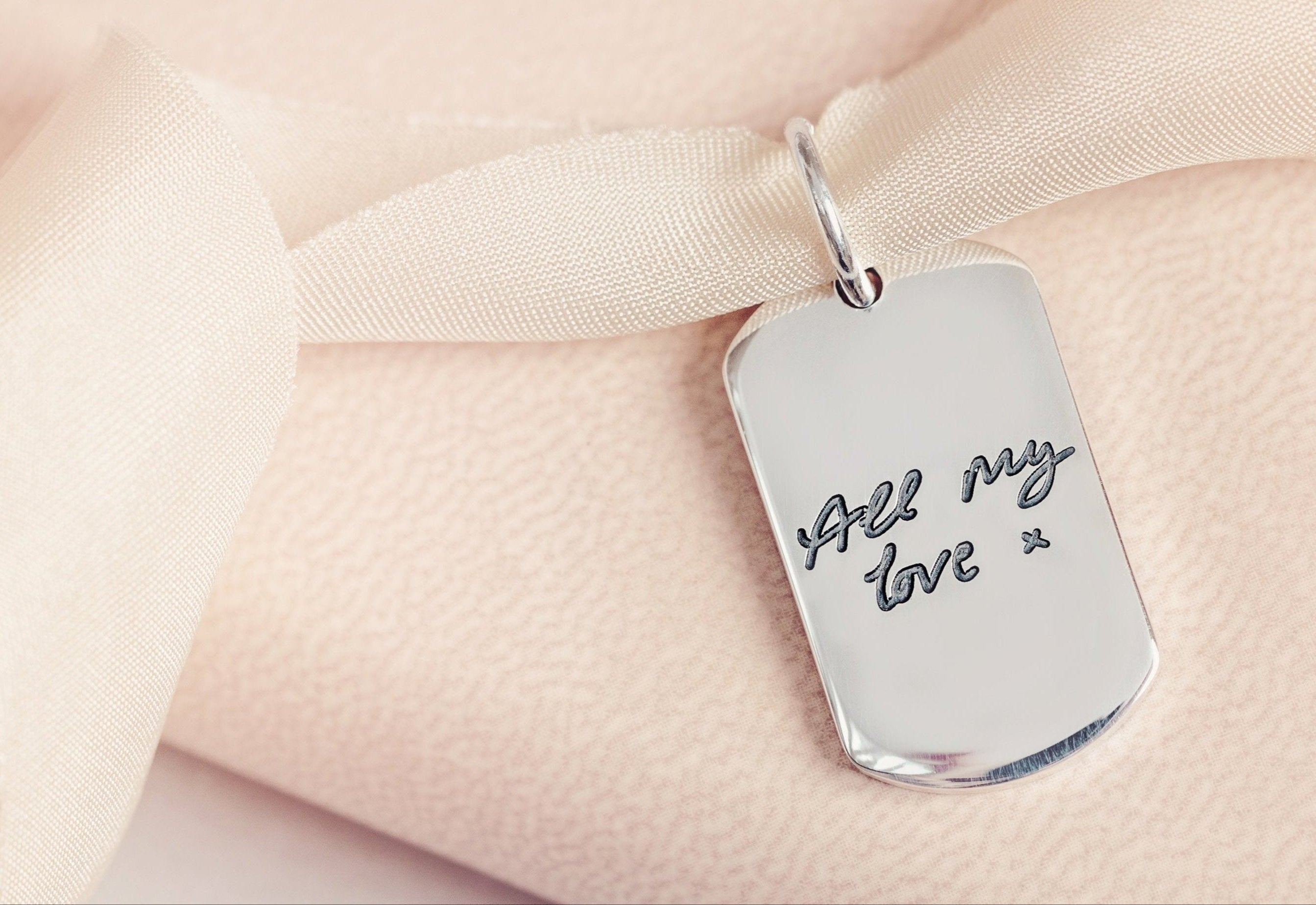 A close up of a dog tag pendant featuring a handwritten message 'All my love x' on a light pink ribbon 