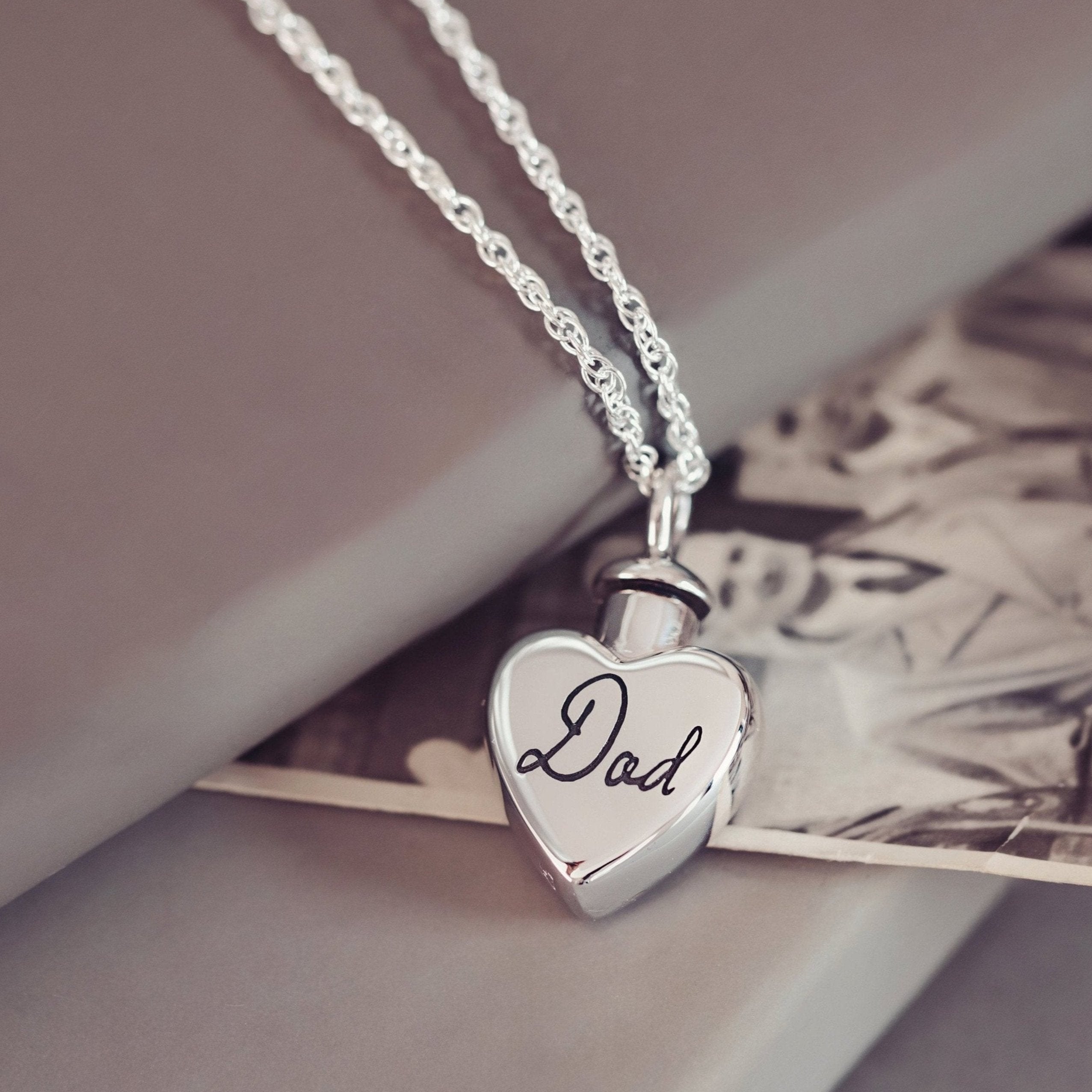 Close up of a silver heart shaped cremation ashes locket engraved with the word 'Dad' in delicate handwriting. The locket rests on top of a vintage black and white photograph