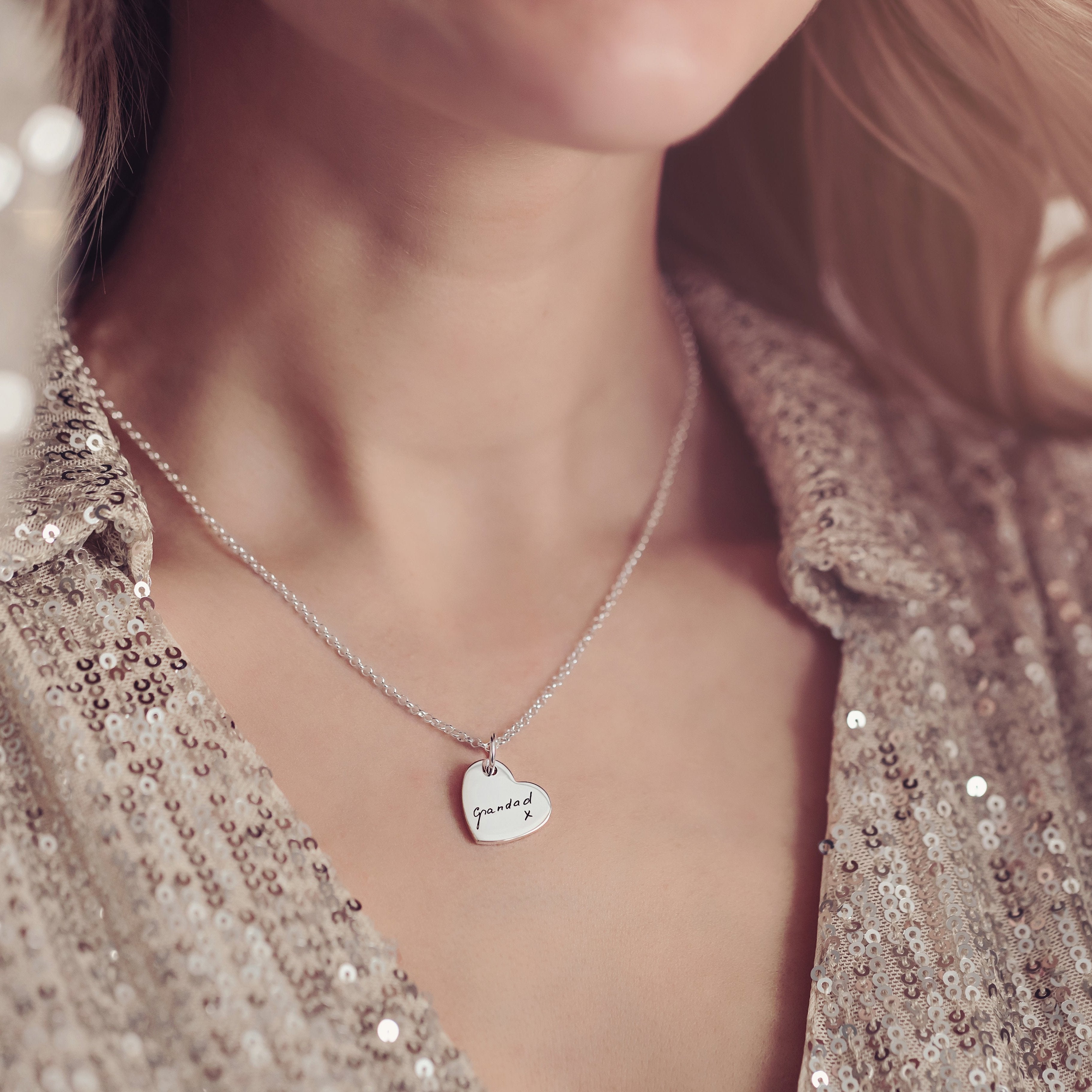 Close up of a silver heart shaped necklace engraved with the handwritten word 'grandad' and a small 'x' beneath it. The necklace is worn around the neck, paired with a sparkly sequin top