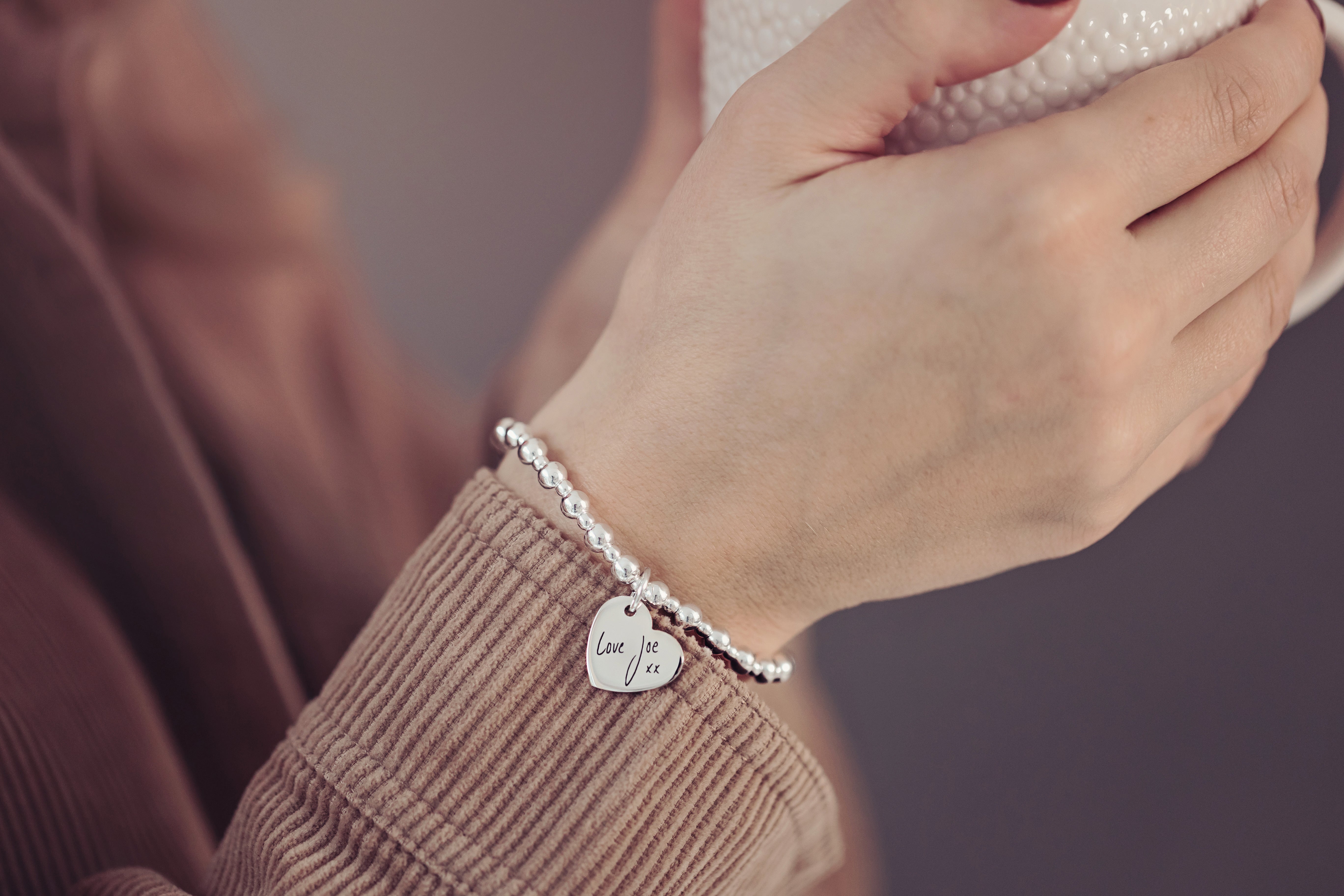 Close up of a silver beaded braclet with heart shaped charm, engraved with handwritten words 'Love Joe xx'. The bracelet is worn on a person's wrist who is holding a white textured cup