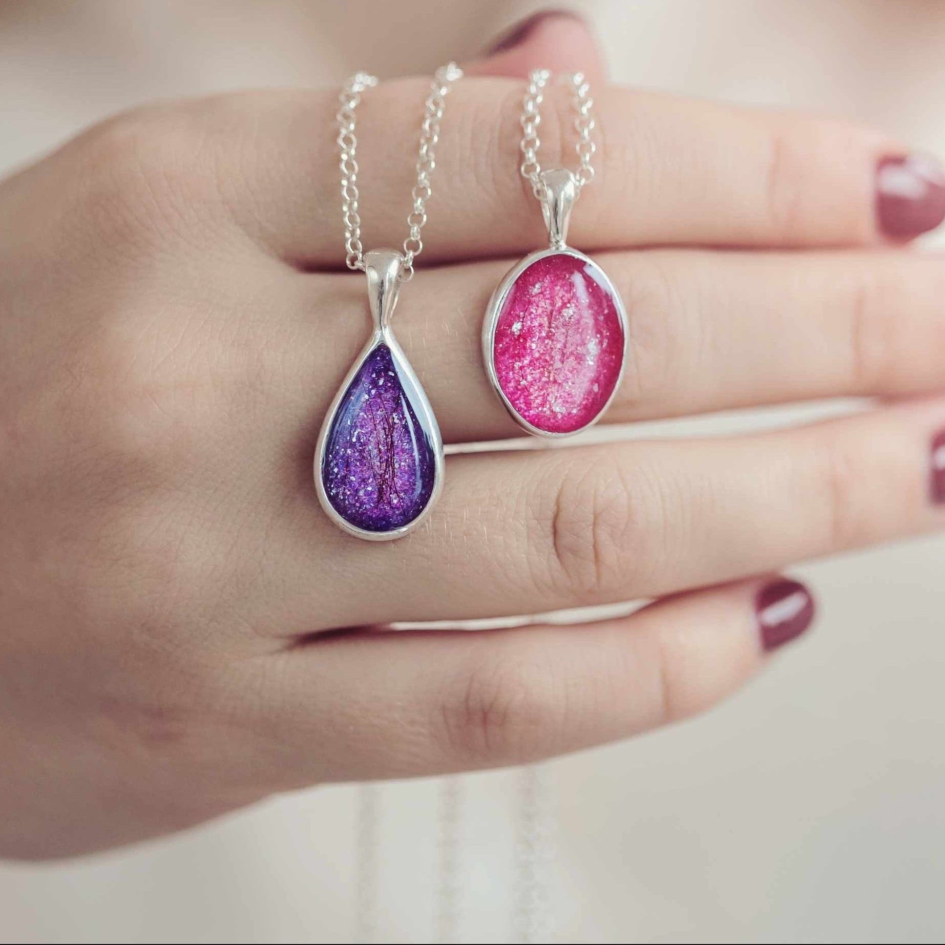 A close-up of a hand holding two memorial hair necklaces, each featuring a sterling silver pendant with a teardrop and oval shape. The pendants are filled with resin containing hair and shimmering crystal details, one in lavender and the other in coral. 