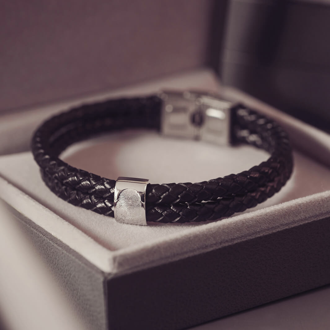 Black braided leather bracelet featuring a silver charm engraved with a fingerprint, displayed in a grey jewellery box