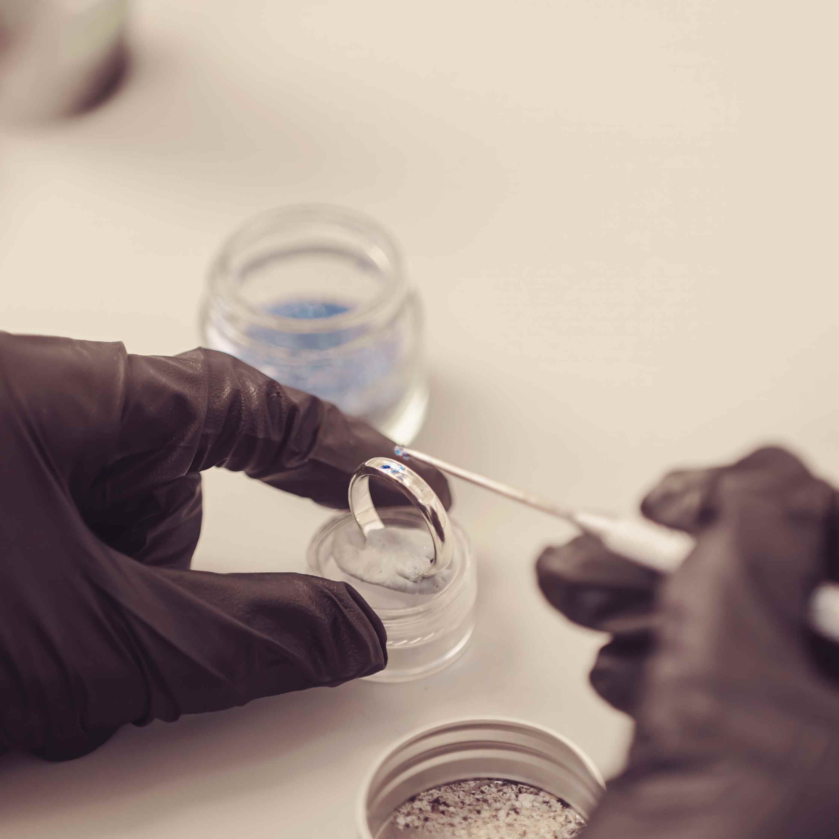 a close up of someone wearing black protective gloves creating a sterling silver womens ring band, carefully adding the ashes and blue crystal into the ring band using a pointed metal tool