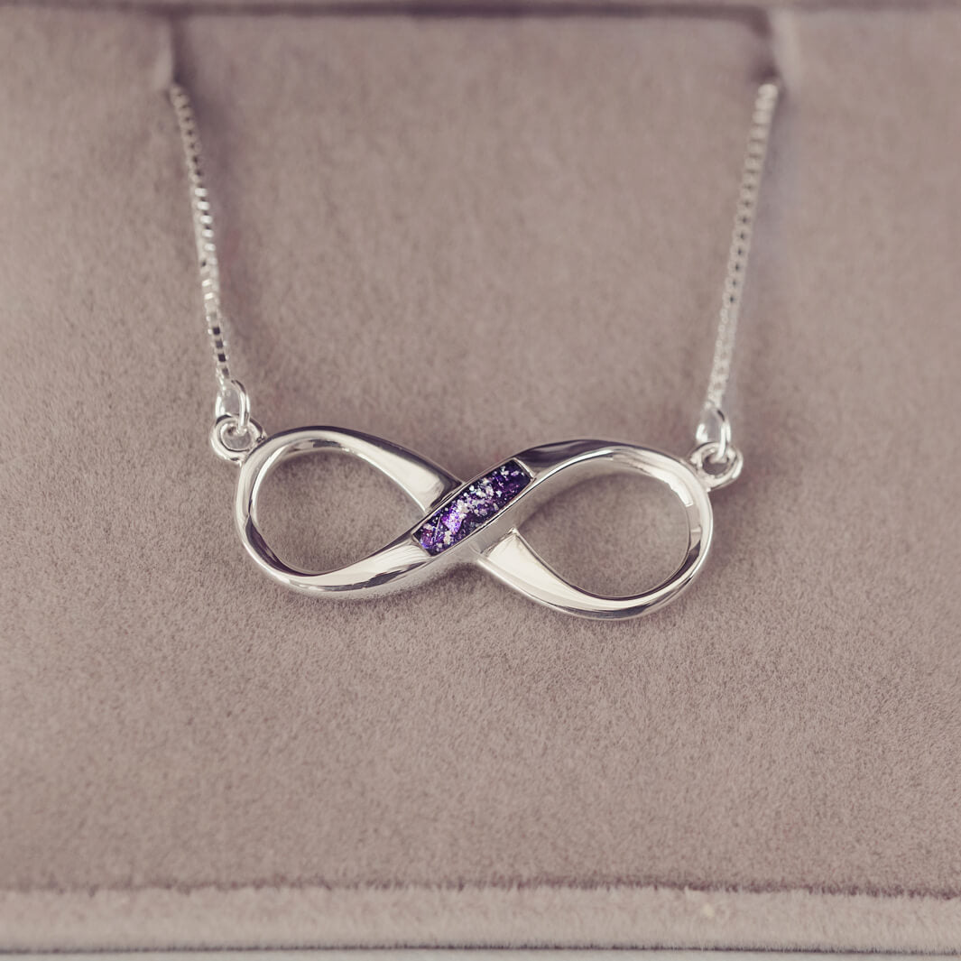 Sterling silver infinity necklace with a central section filled with memorial ashes and purple crushed crystals, displayed on a beige fabric background