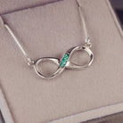 Sterling silver infinity necklace with a central section filled with memorial ashes and green crushed crystals, displayed on a beige fabric background