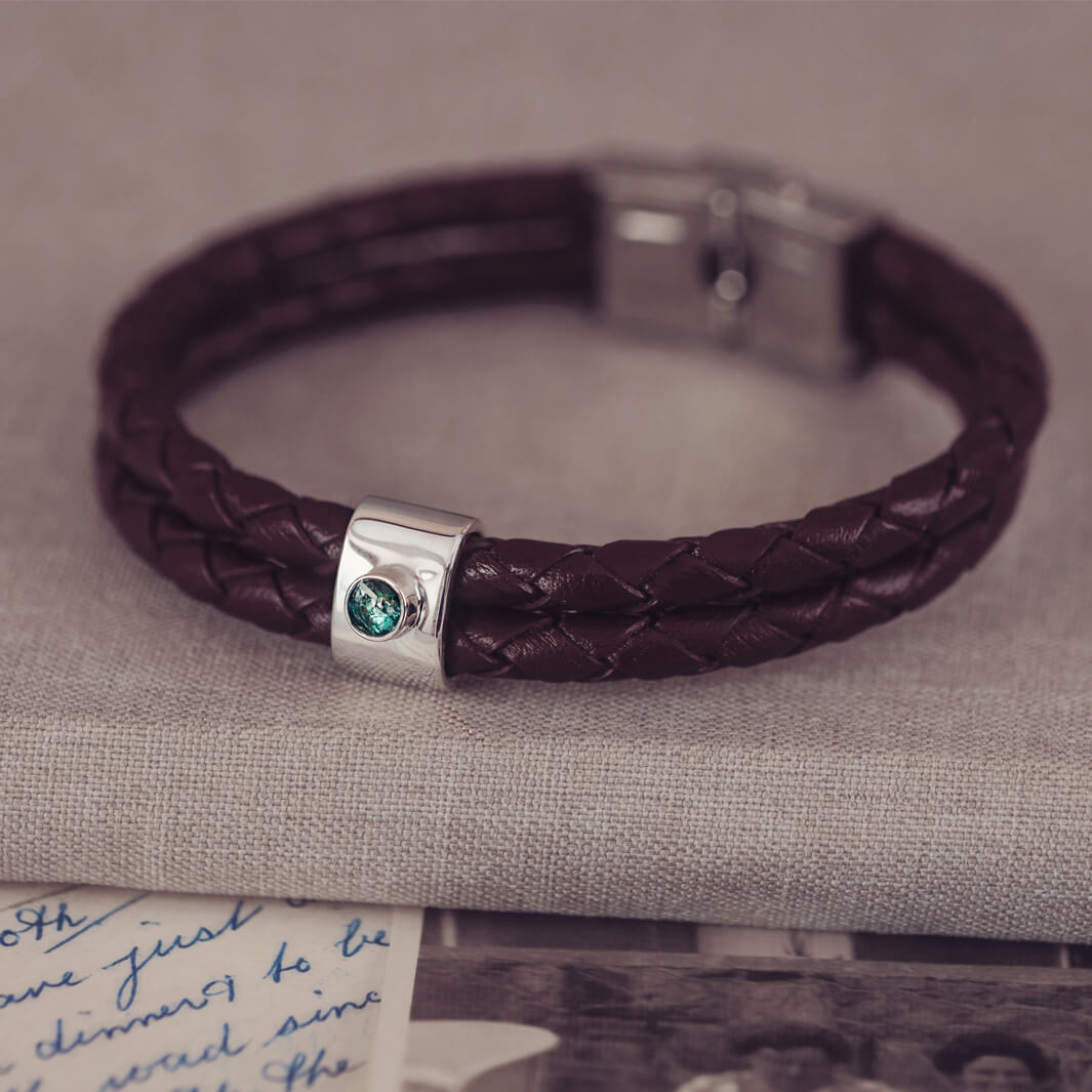 Brown braided leather bracelet featuring a silver charm with a small green memorial stone set in the centre, displayed on a grey surface.