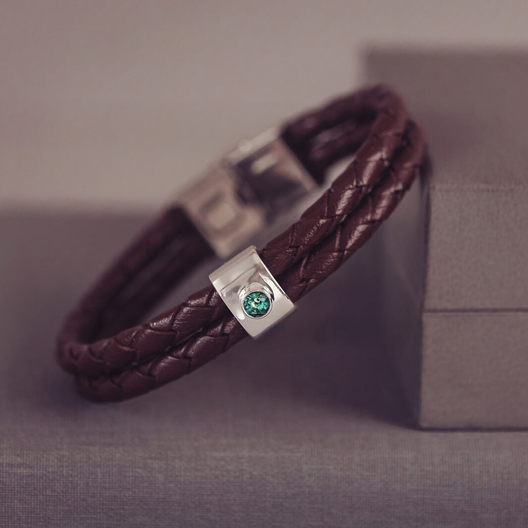 Brown braided leather bracelet featuring a silver charm with a small green memorial stone set in the centre, displayed on a grey surface.