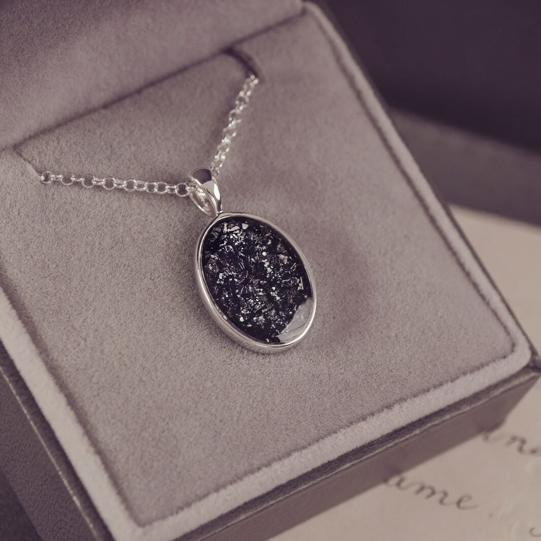 Sterling silver oval necklace with a central section filled with memorial ashes and black crushed crystals, displayed in a grey jewellery box