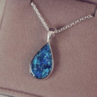 Sterling silver teardrop necklace with a central section filled with memorial ashes and blue crushed crystals, displayed on a grey fabric background