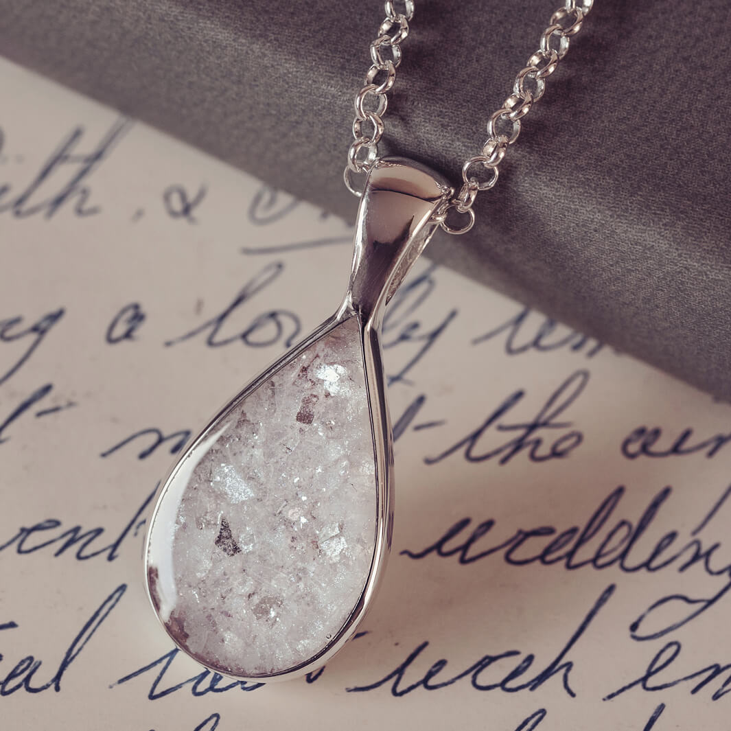 Sterling silver teardrop necklace with a central section filled with memorial ashes and white and clear crushed crystals, displayed on a white paper background with a handwritten message in blue ink