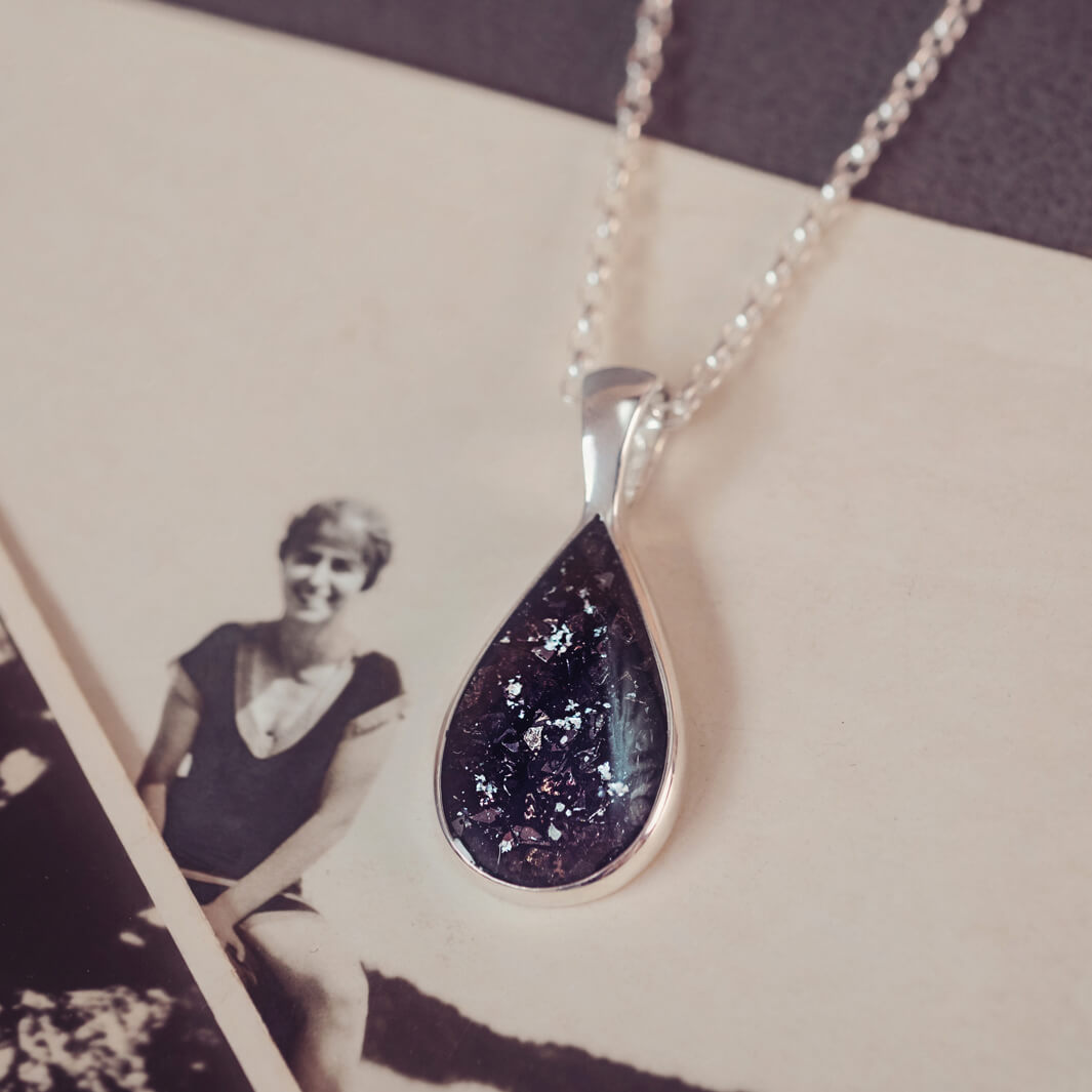 Sterling silver teardrop necklace with a central section filled with memorial ashes and black crushed crystals, displayed on a background of an old photograph of a woman in a swimming cap