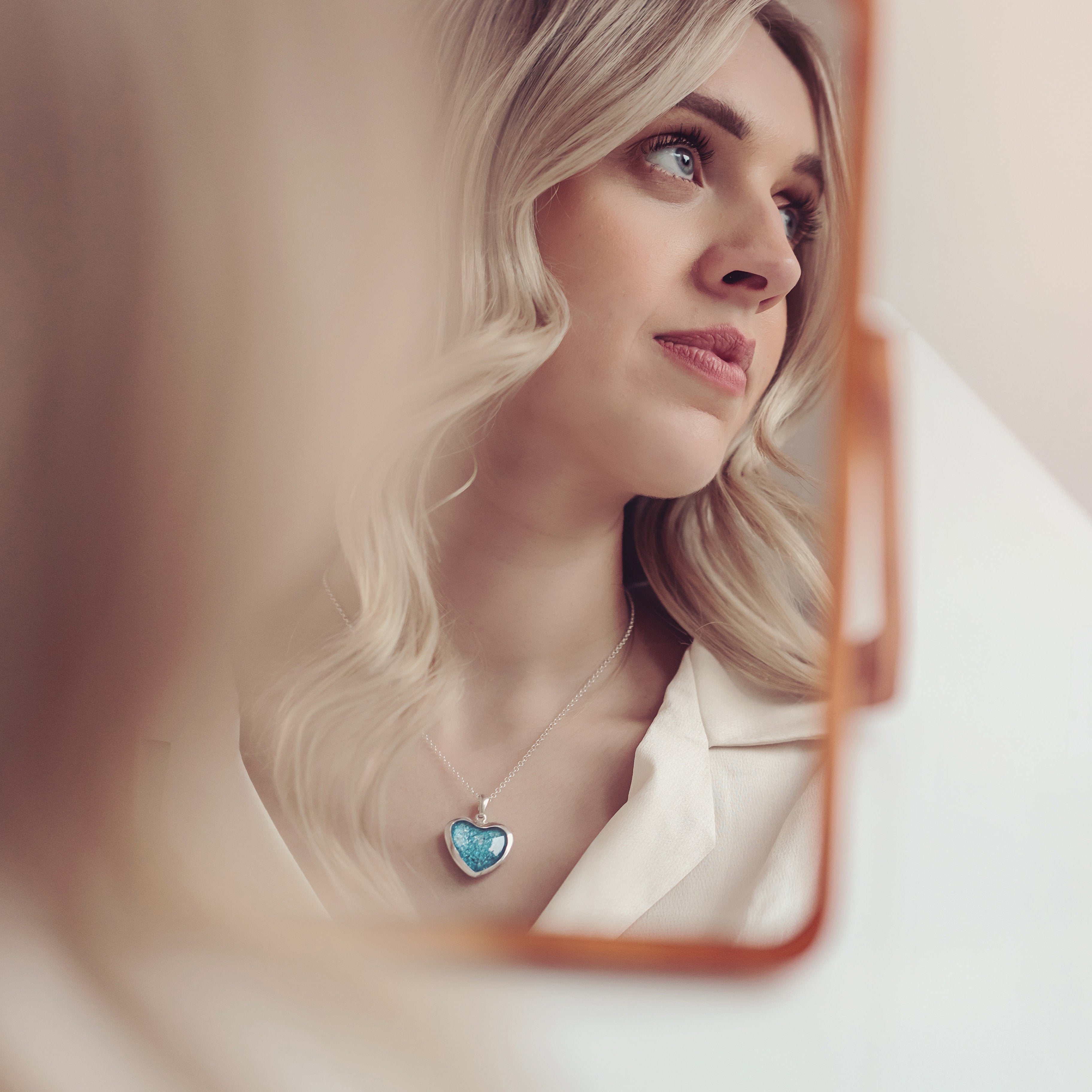 Woman with long blonde hair looking into mirror, wearing a white shirt and a blue heart memorial hair necklace