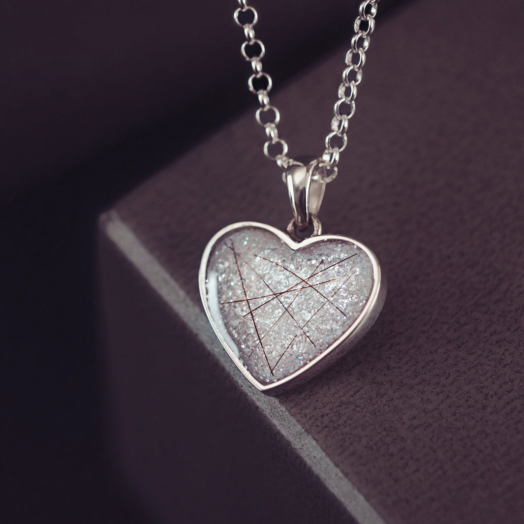 A silver heart shaped necklace charm, on a silver chain, filled with shite glitter, strands of dark hair, and resin.