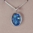 Sterling silver oval necklace with a central section filled with memorial ashes and blue crushed crystals, displayed on a grey fabric background