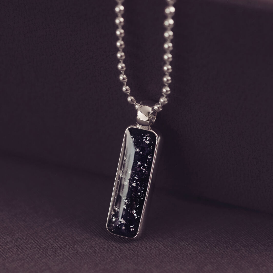 A sophisticated Mens silver necklace featuring a rectangular pendant filled with shimmering black crystal and resin, delicately layered with cremation ashes, showcased against a dark background on a silver ball chain