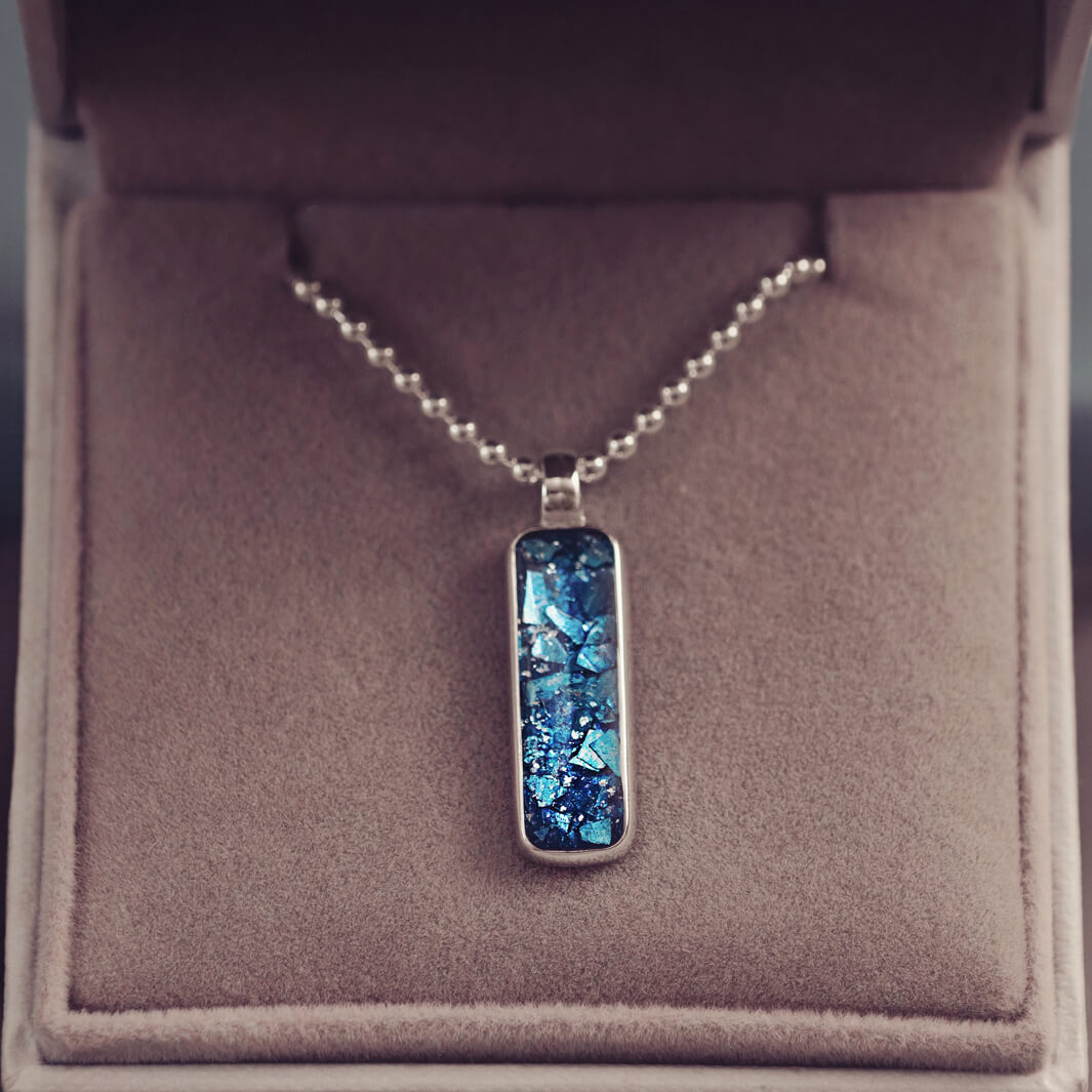 A sophisticated Mens silver necklace featuring a rectangular pendant filled with shimmering blue crystal and resin, delicately layered with cremation ashes, showcased against a dark background on a silver ball chain