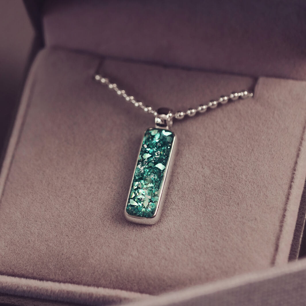 A sophisticated Mens silver necklace featuring a rectangular pendant filled with shimmering green crystal and resin, delicately layered with cremation ashes, showcased against a dark background on a silver ball chain