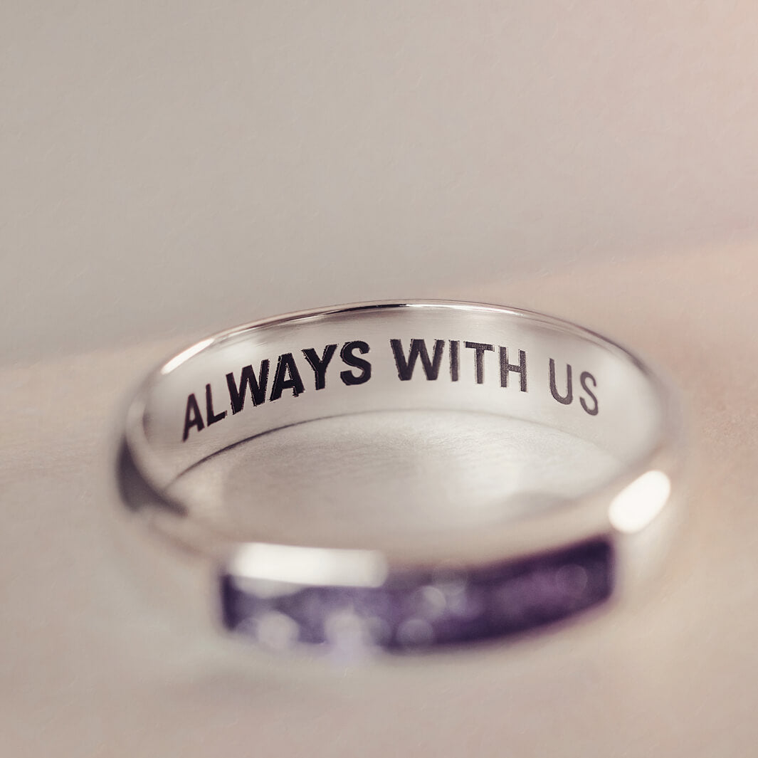 Sterling silver Mens Band Ring with a central section filled with memorial ashes and purple crushed crystals, displayed on a blurred pale pink background. The ring has the message  'ALWAYS WITH US' engraved into the inside of the band in black