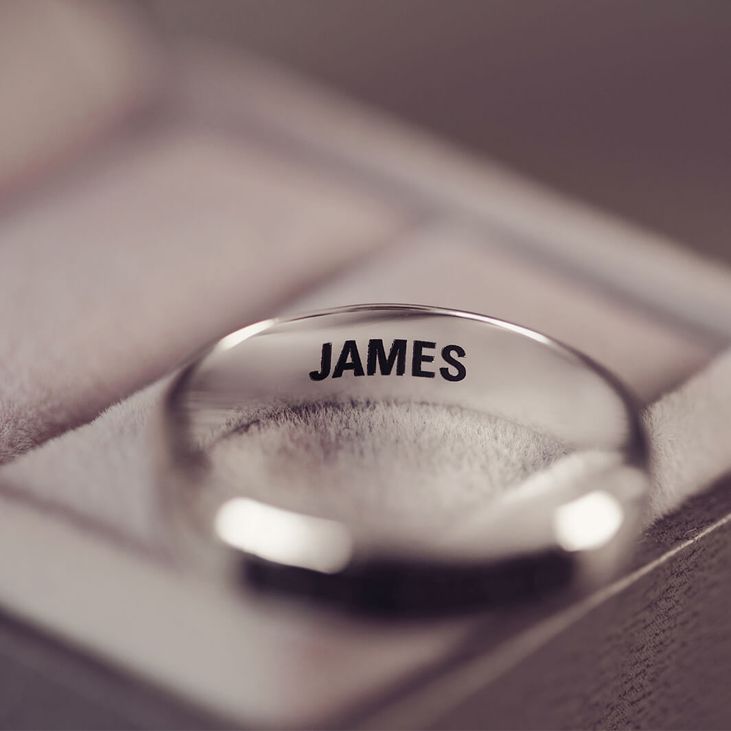 Sterling silver Mens Band Ring with a central section filled with memorial ashes and black crushed crystals, displayed on a blurred grey background. The ring has the message 'JAMES' engraved into the inside of the ring band in black