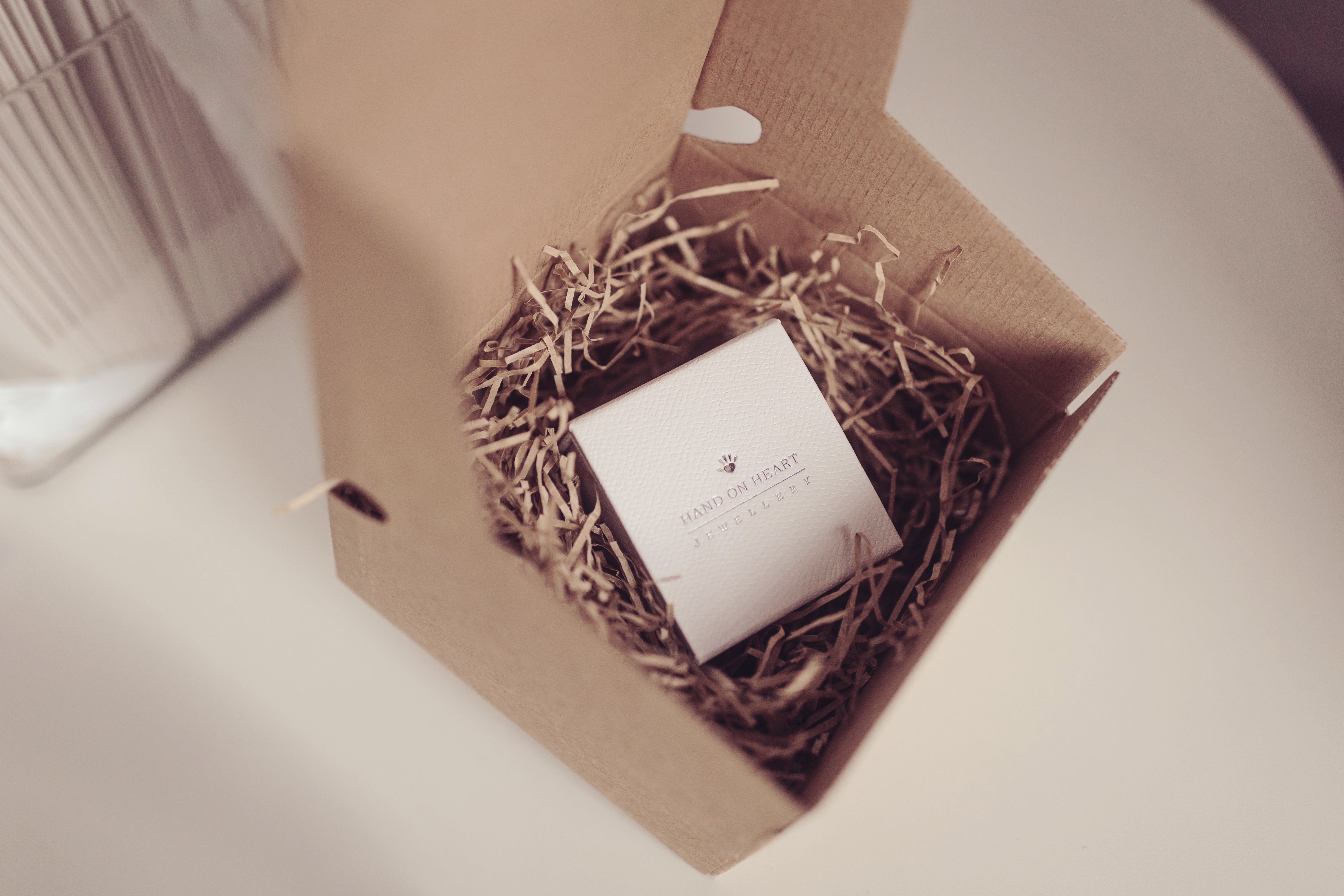 A white jewellery box nestled in a box of brown shredded paper placed on a table