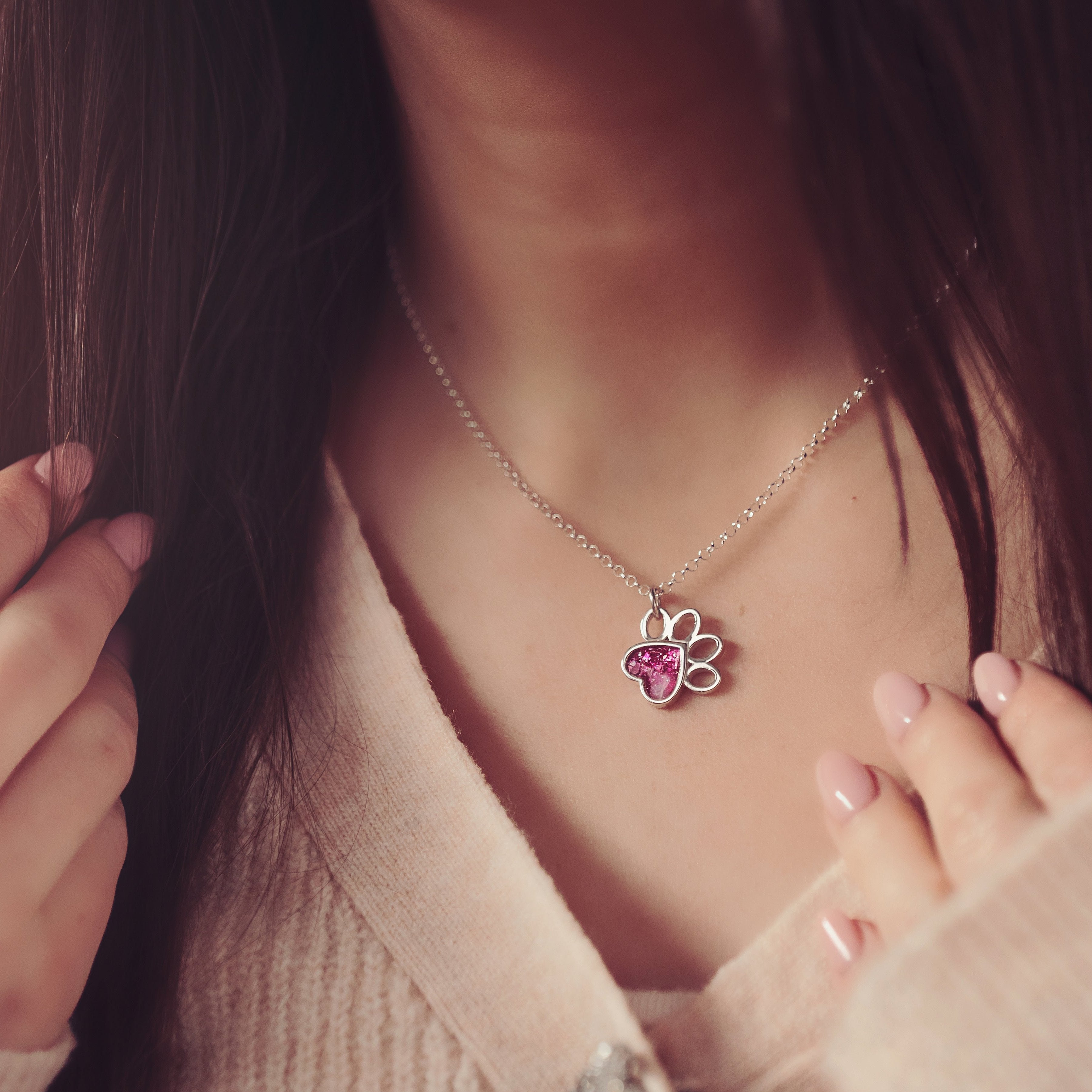 Close up of a silver necklace with a pink paw-shaped pendant made with cremation ashes, worn by a woman with long dark hair, gently holding a strand of her hair