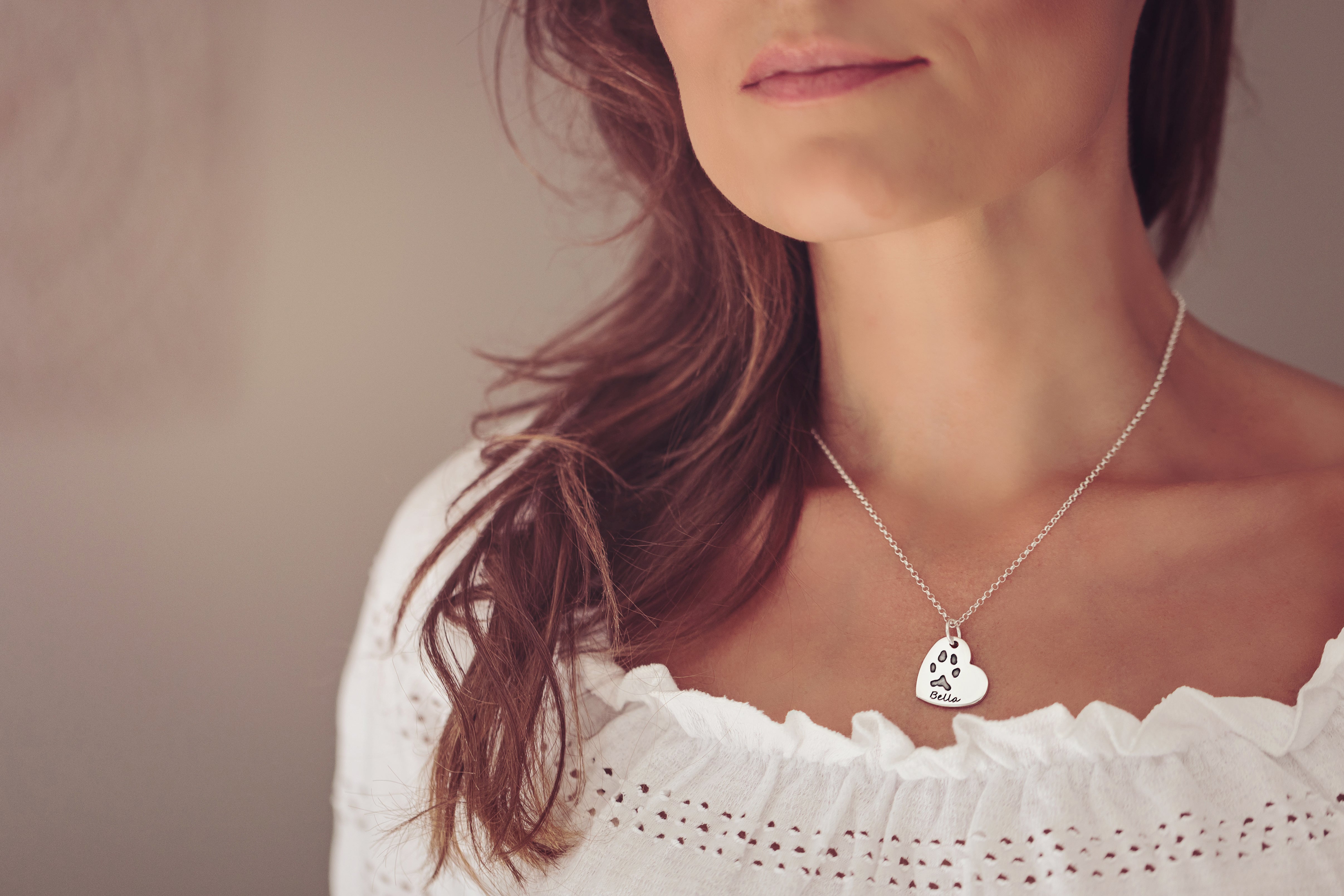 A woman wearing a white top and a heart necklace featuring a pawprint and name 'Bella' engraved