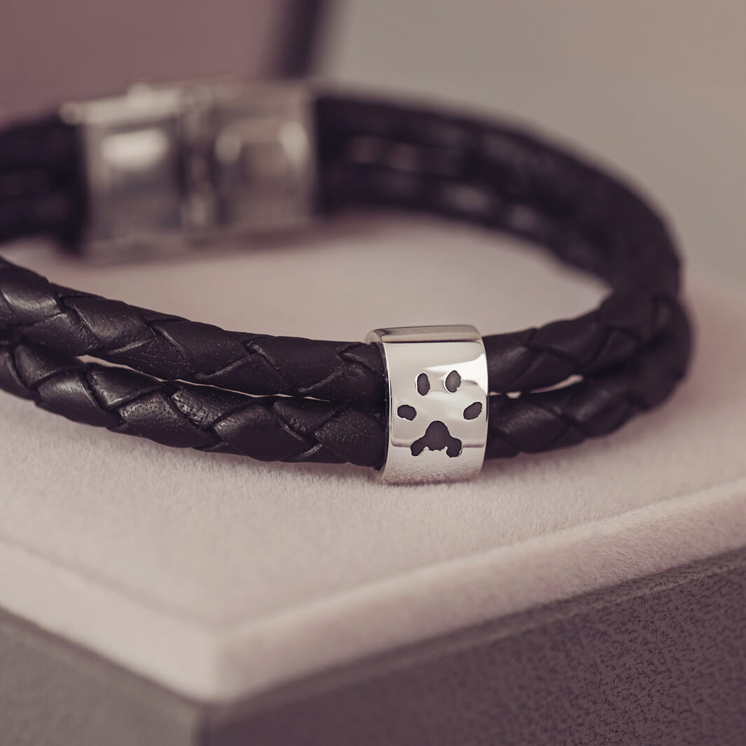 Black braided leather bracelet featuring a silver charm engraved with a black pawprint, displayed on a soft grey background.