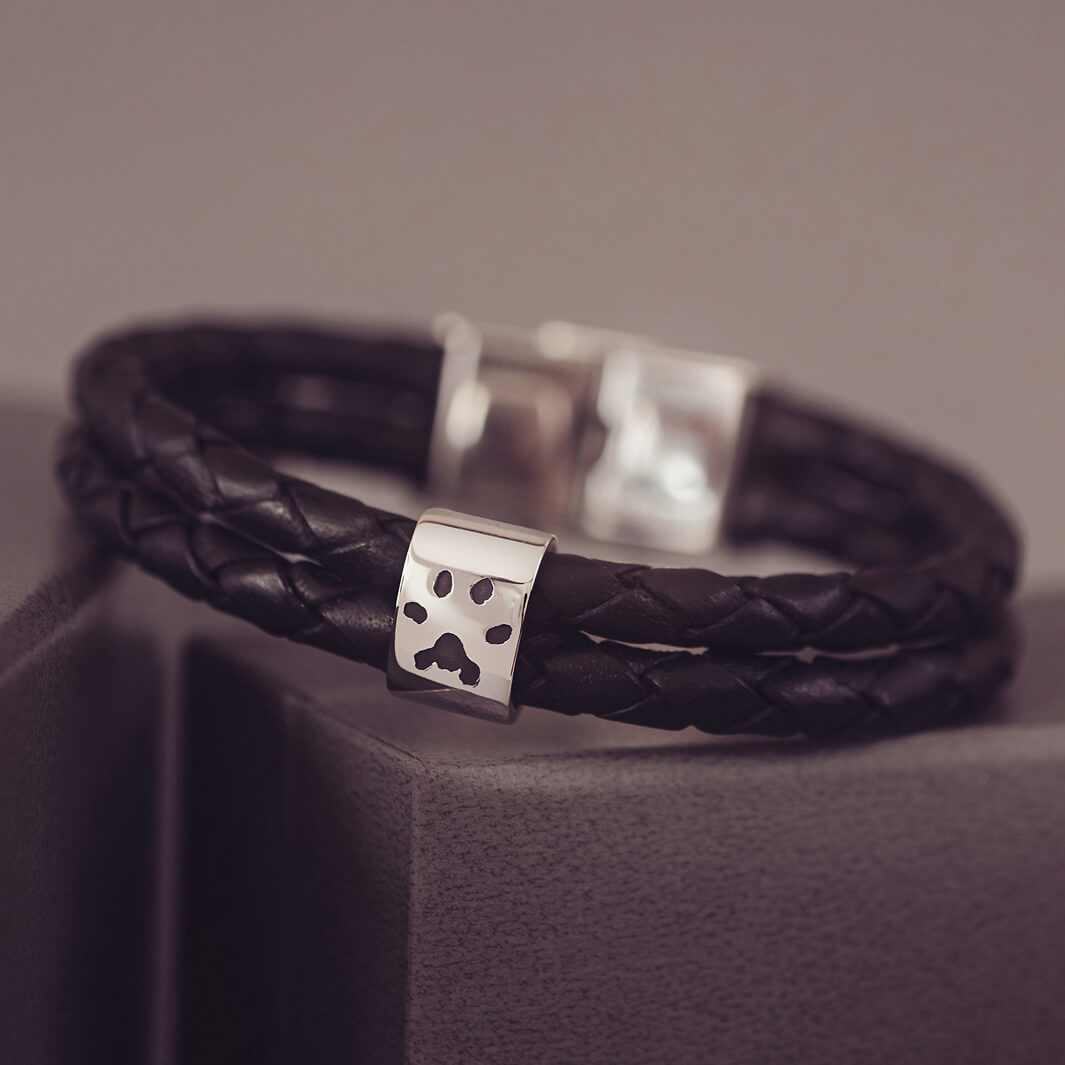 Black braided leather bracelet featuring a silver charm engraved with a black pawprint, displayed on a soft grey background.