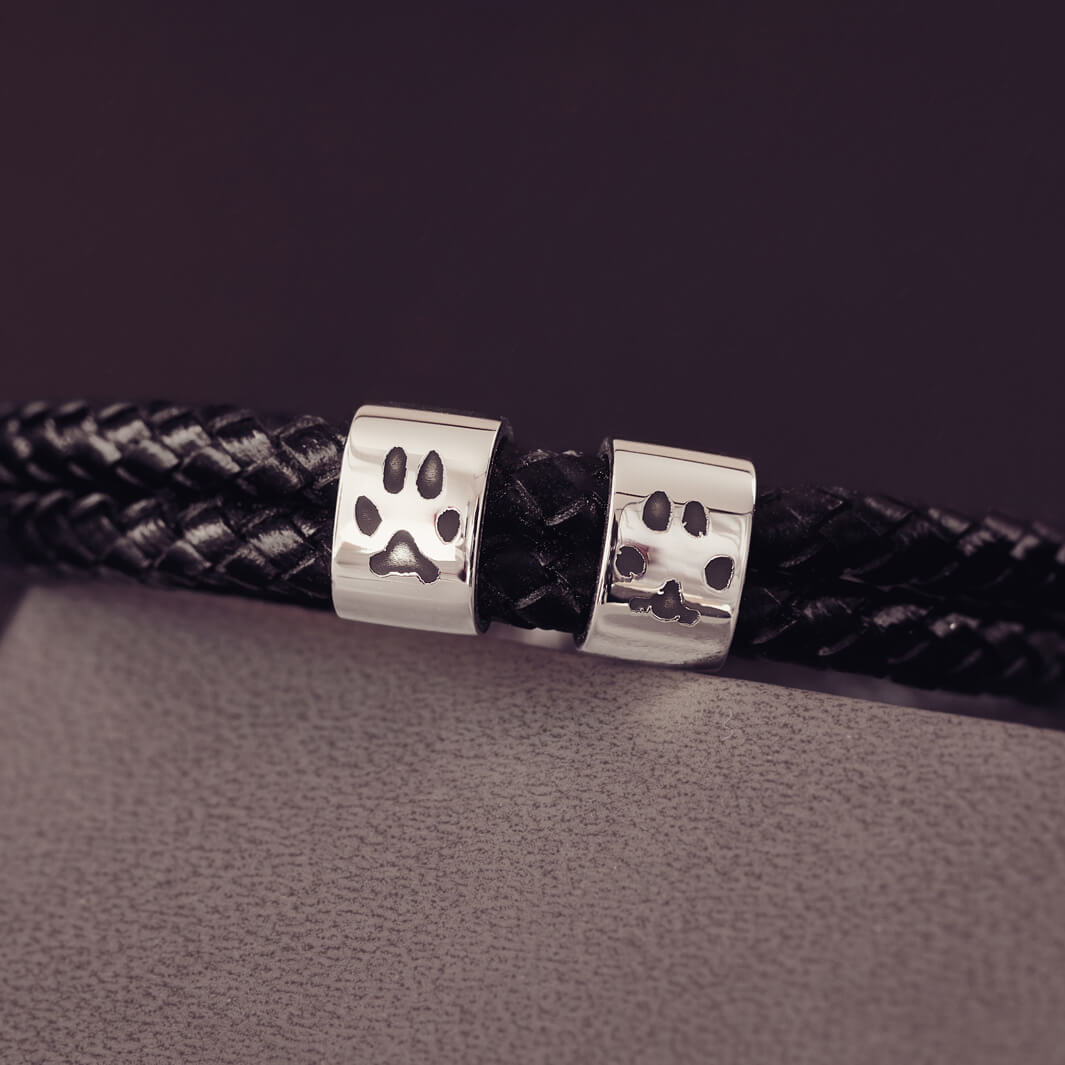 Black braided leather bracelet featuring two sterling silver charms, each engraved with a black pawprint, displayed on a soft grey background.