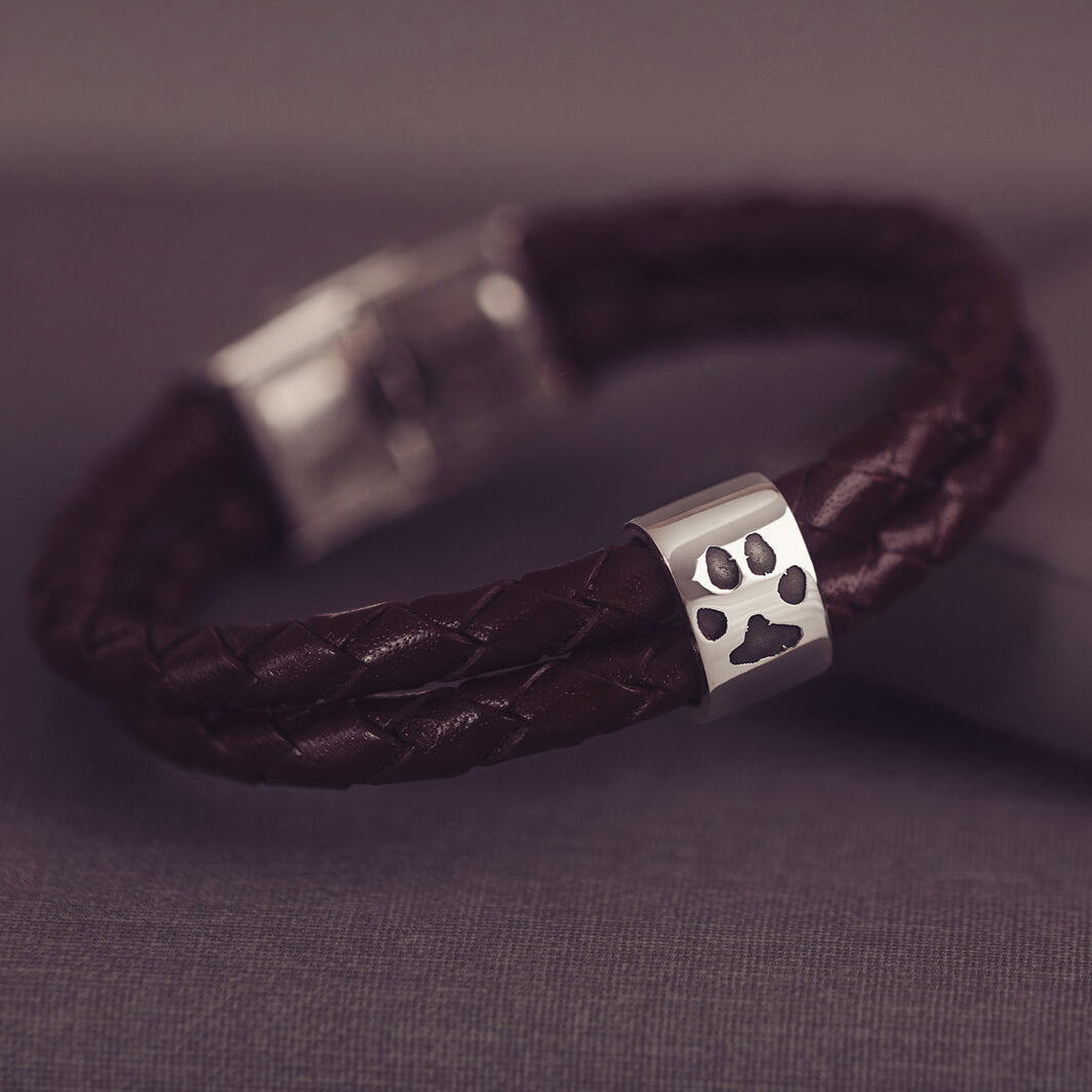 Brown braided leather bracelet featuring a silver charm engraved with a black pawprint, displayed on a dark grey background.