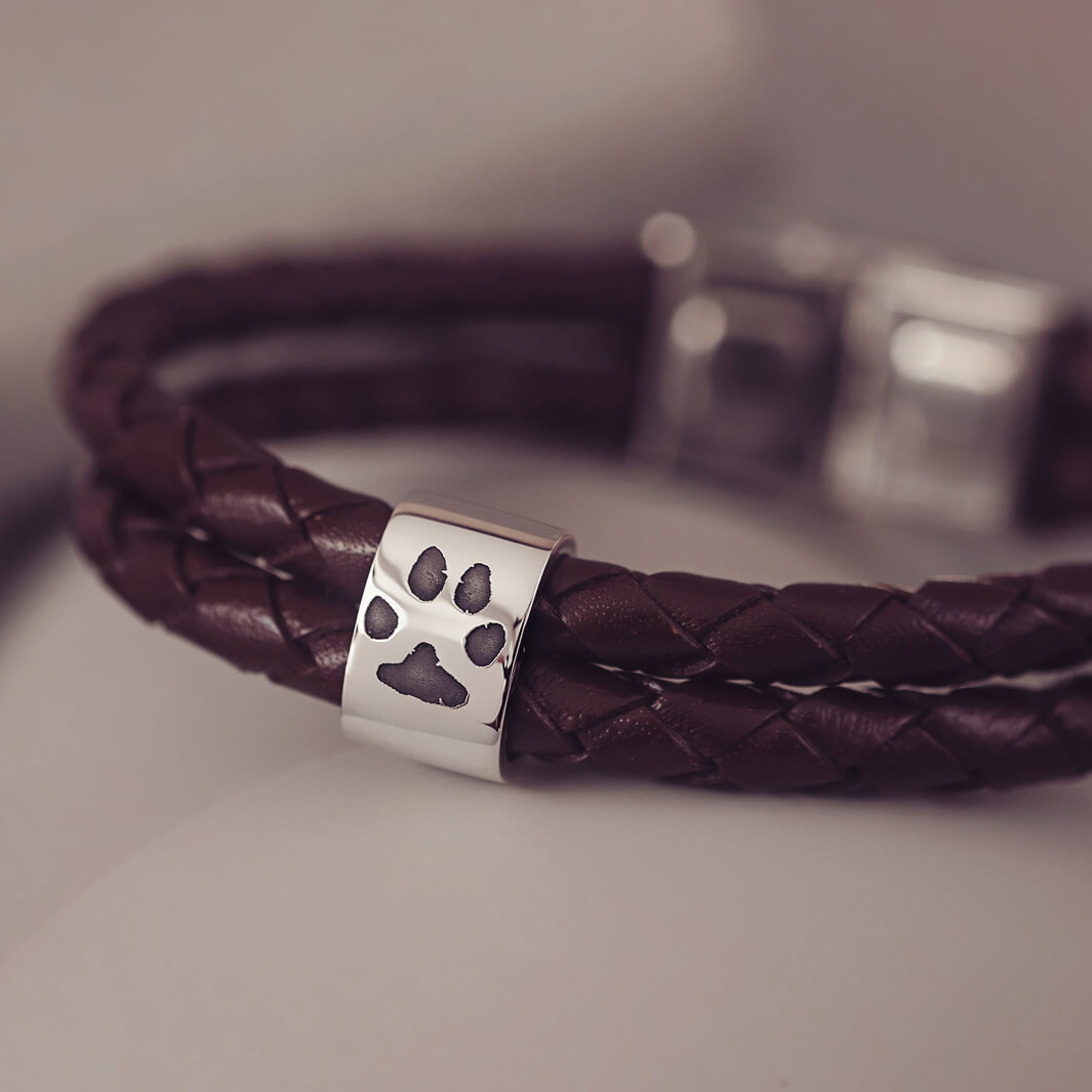 Brown braided leather bracelet featuring a silver charm engraved with a black pawprint, displayed on a soft grey background.