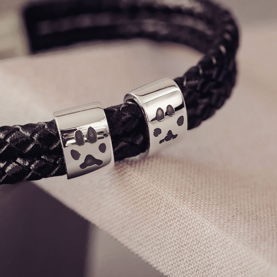 Black braided leather bracelet featuring two sterling silver charms, each engraved with a black pawprint, displayed on a soft grey background.