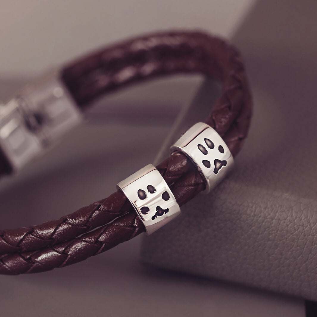 Brown braided leather bracelet featuring two sterling silver charms, each engraved with a black pawprint, displayed on a soft grey background.