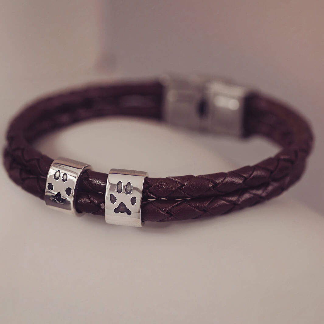 Black braided leather bracelet featuring two sterling silver charms, each engraved with a black pawprint, displayed on a soft grey background.