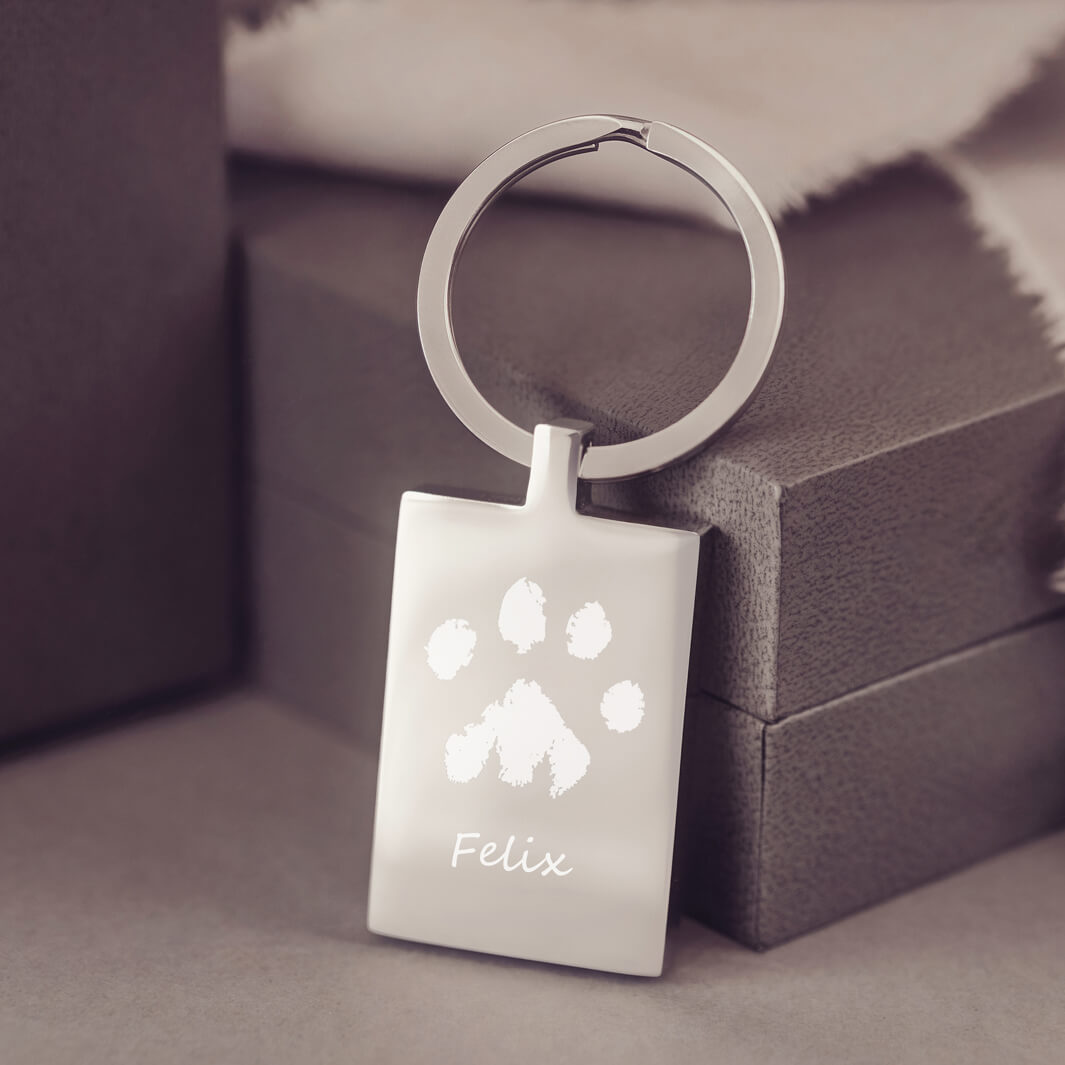 A rectangular keyring, engraved with a pawprint and name, resting against a dark grey jewellery box