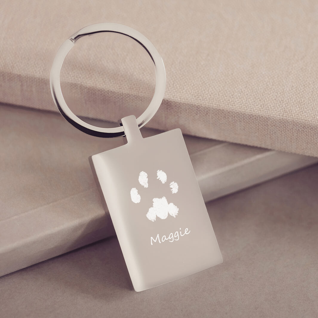 A rectangular stainless steel keyring, engraved with one pawprint and name, leaning on a dusky pink fabric background