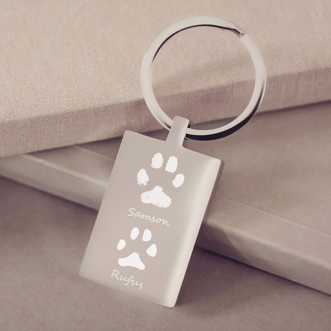 A rectangular stainless steel keyring, engraved with two pawprints and names, leaning on a dusky pink fabric background