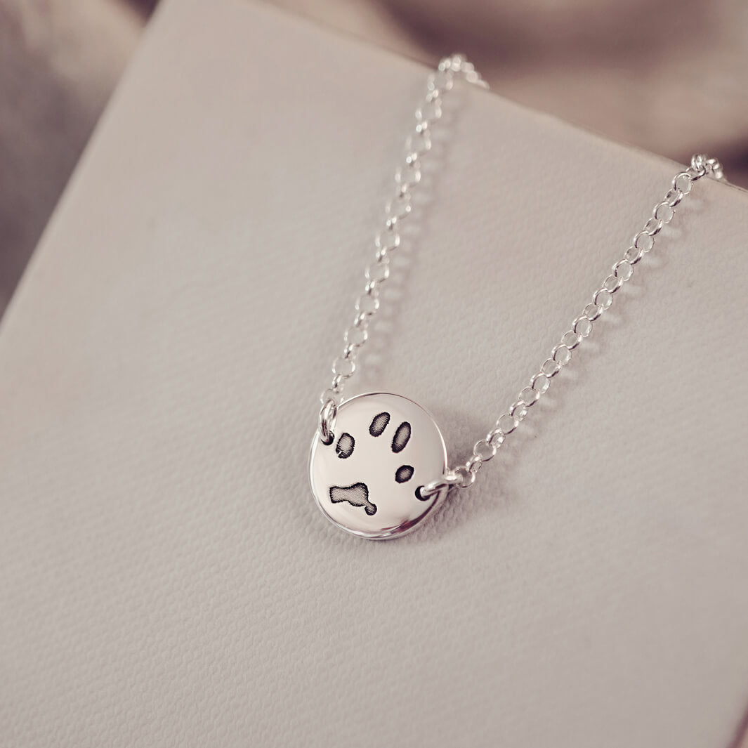 A round silver pendant bracelet featuring a detailed engraved pawprint, displayed on a delicate silver chain. The necklace rests in an elegant, soft velvet jewellery box