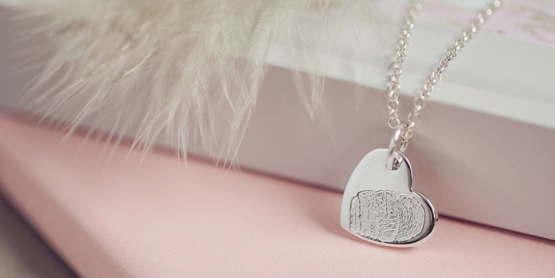 A close up of a fingerprint necklace resting on a soft pink surface with a grey feather in the background