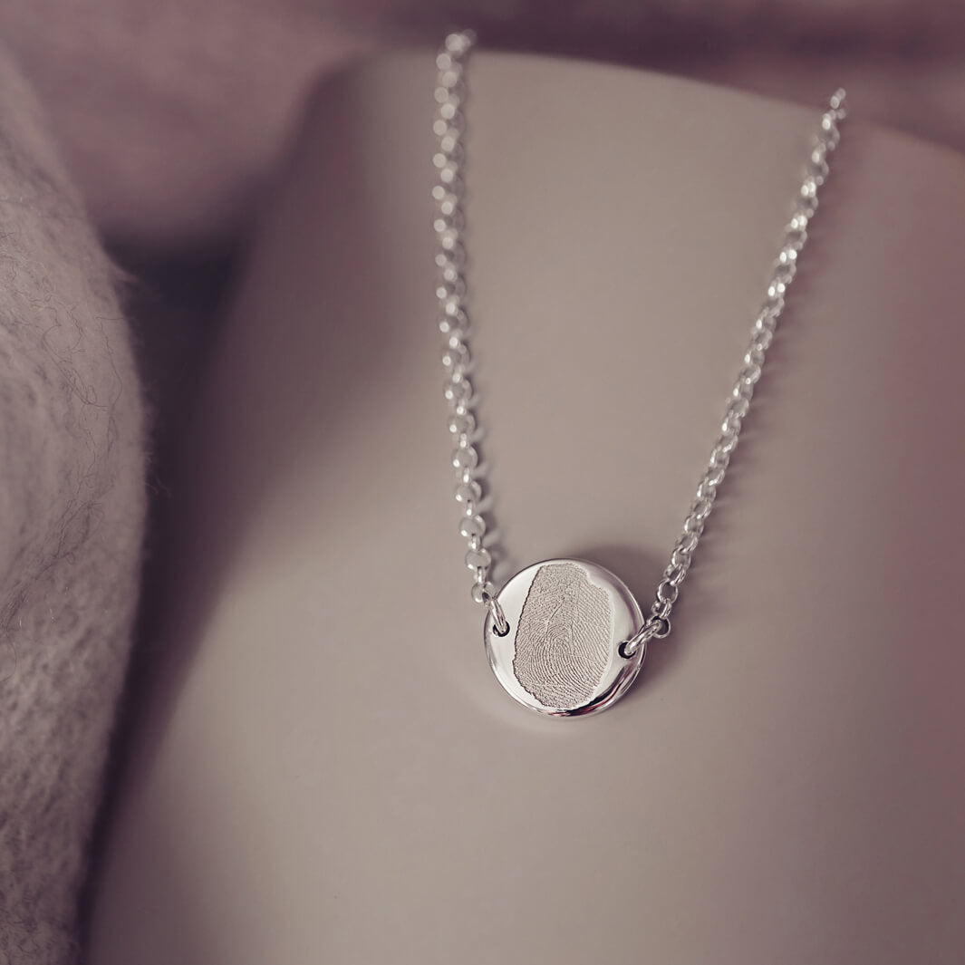 A round silver pendant bracelet featuring a detailed imprinted fingerprint, displayed on a delicate silver chain. The necklace rests in an elegant, soft velvet jewellery box