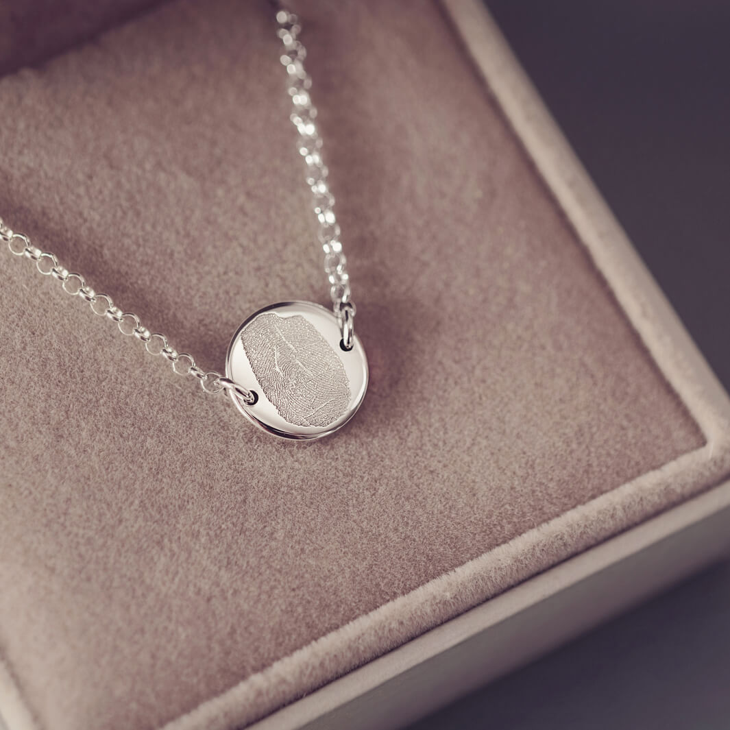 A round silver pendant bracelet featuring a detailed imprinted fingerprint, displayed on a delicate silver chain. The necklace rests in an elegant, soft velvet jewellery box