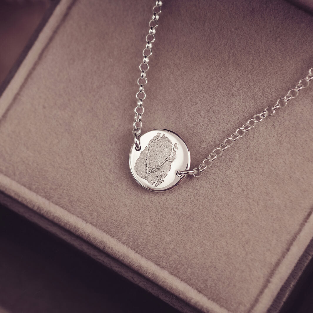 A round silver pendant bracelet featuring a detailed imprinted fingerprint, displayed on a delicate silver chain. The necklace rests in an elegant, soft velvet jewellery box