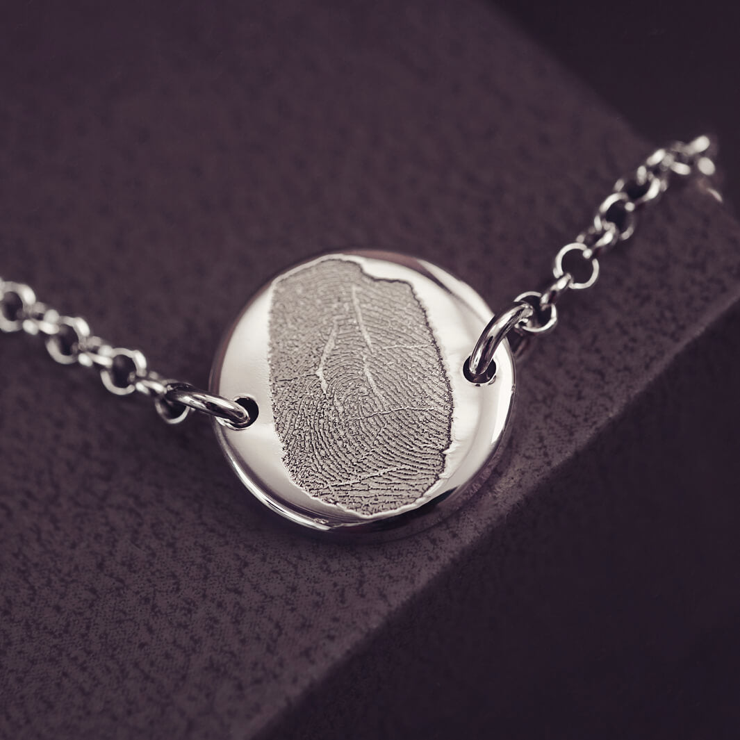 A round silver pendant bracelet featuring a detailed imprinted fingerprint, displayed on a delicate silver chain. The necklace rests in an elegant, soft velvet jewellery box