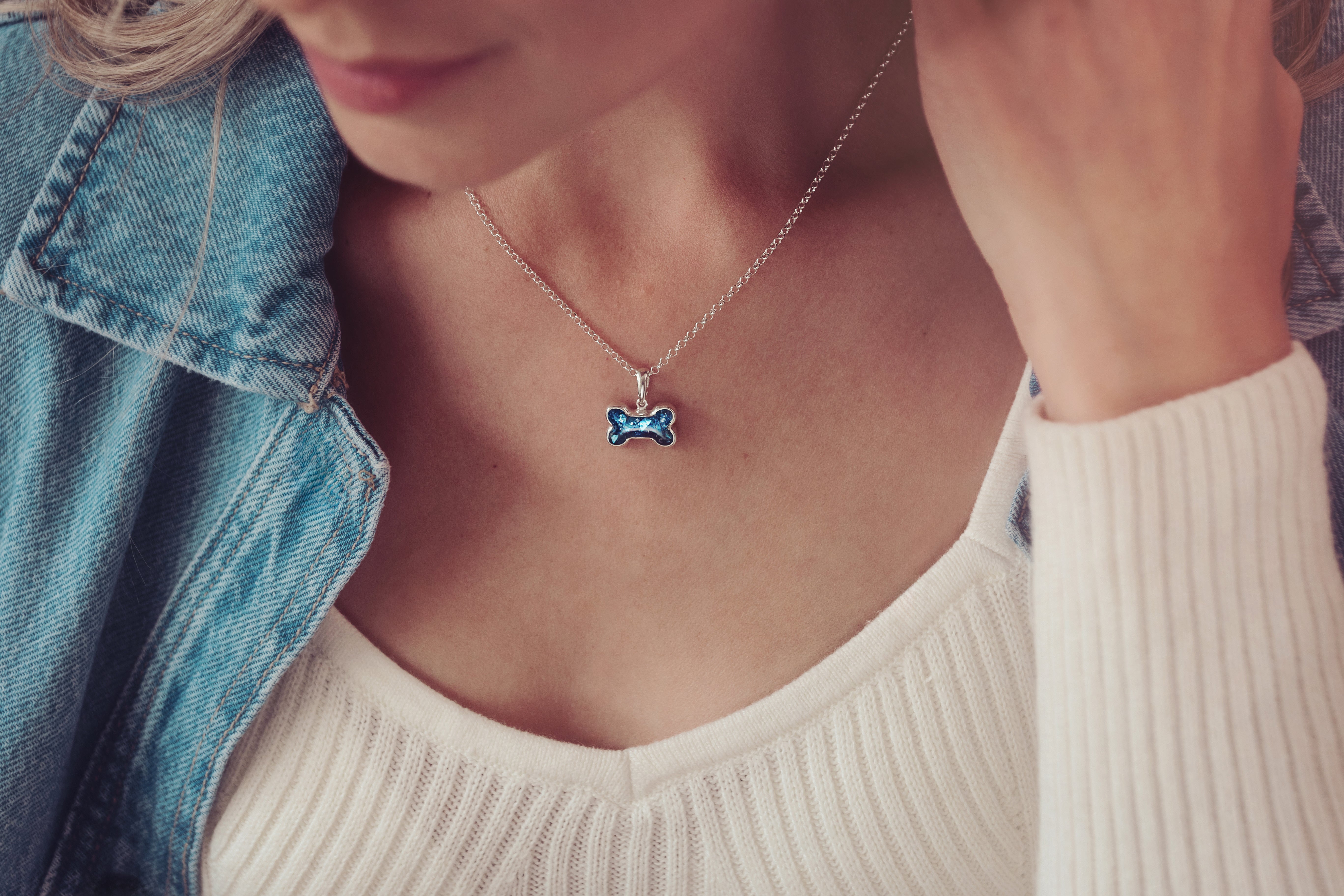 Silver necklace featuring a blue pet tag pendant made with cremation ashes, worn by a woman in white top and denim jacket