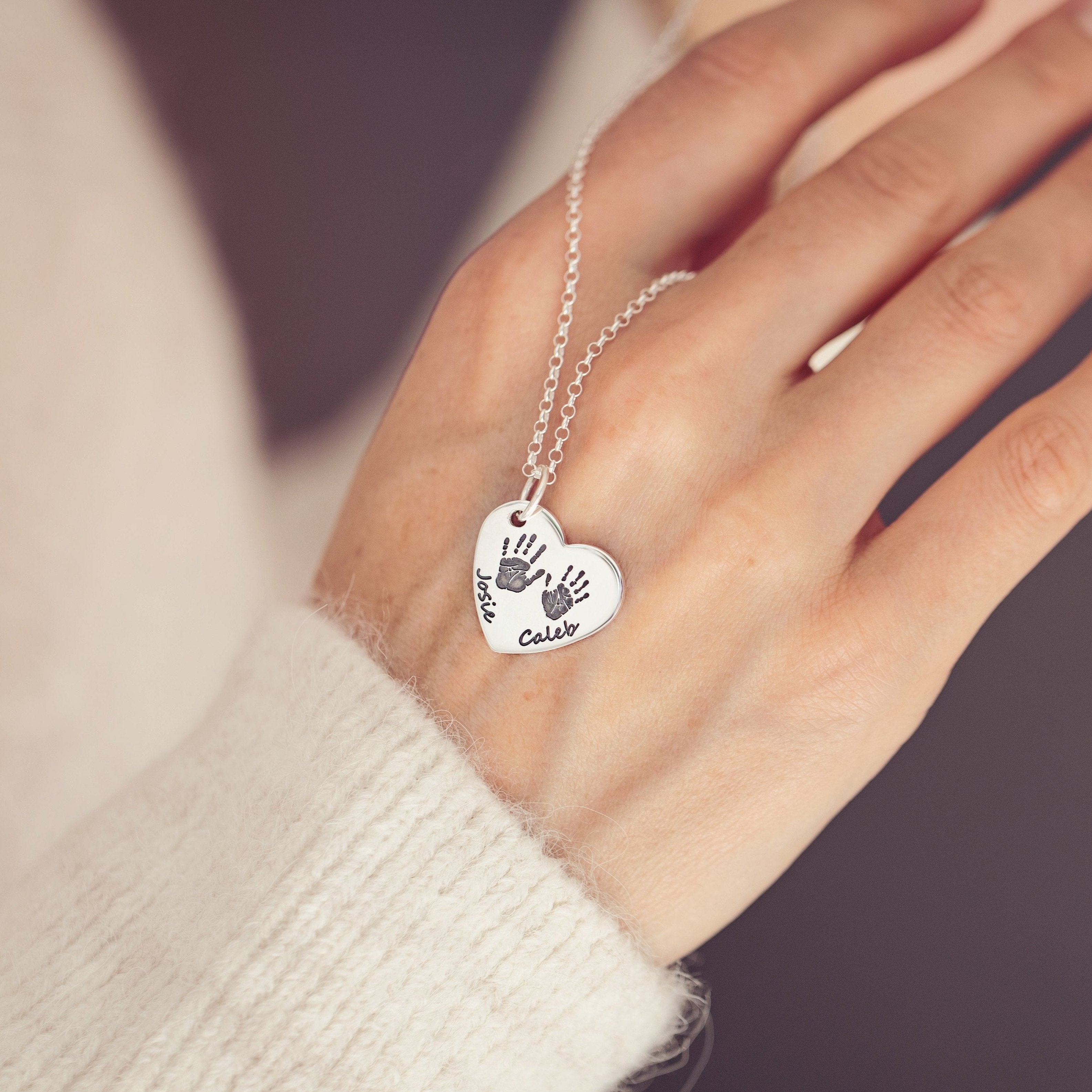 A woman elegantly holds a heart necklace featuring two handprints and names 'Josie' and 'Caleb' engraved