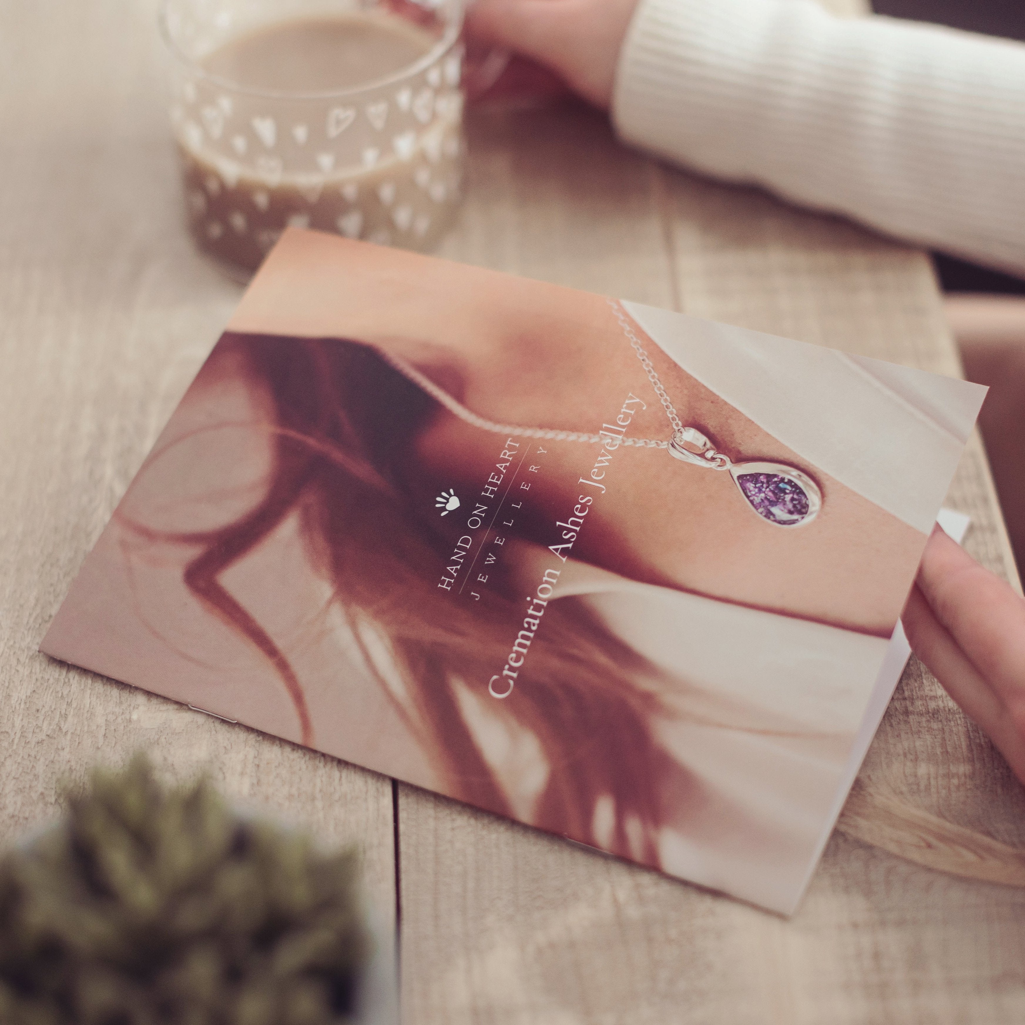 Close up of a woman opening a Cremation Ashes Jewellery brochure at a desk, holding a cup of coffee in her other hand