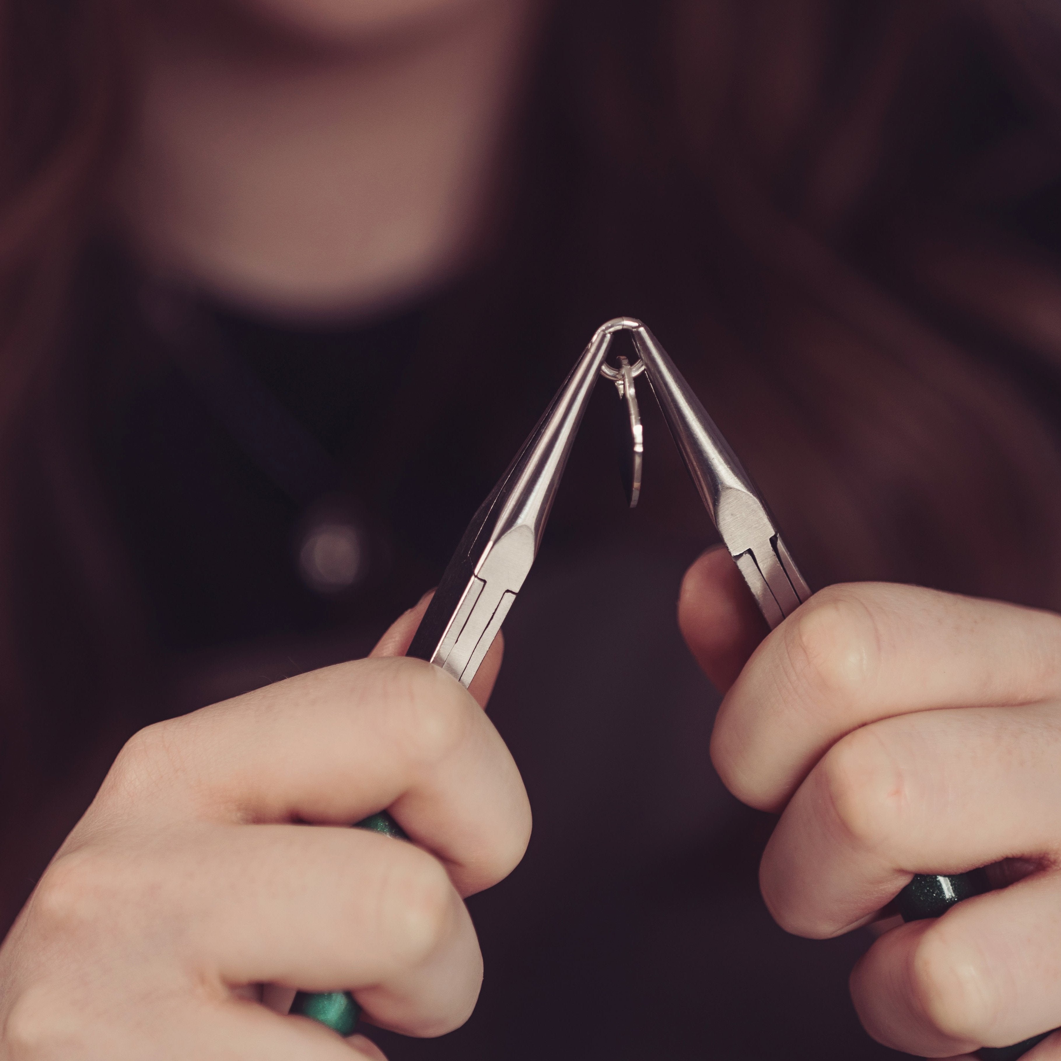 A woman holding a heart pendant with pliers, preparing to create a necklace