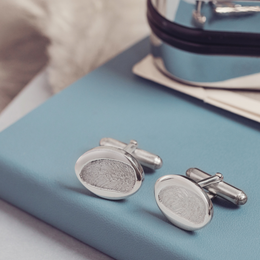 A pair of silver fingerprint cufflinks placed on a blue book. In the background, the mirrored cufflink case is blurred