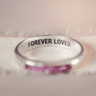 Sterling silver womens ring band with a central section filled with  memorial ashes and pink crushed crystals, displayed on a blurred fabric background. The ring has the message 'Forever Loved' engraved into the inside of the ring band, in black